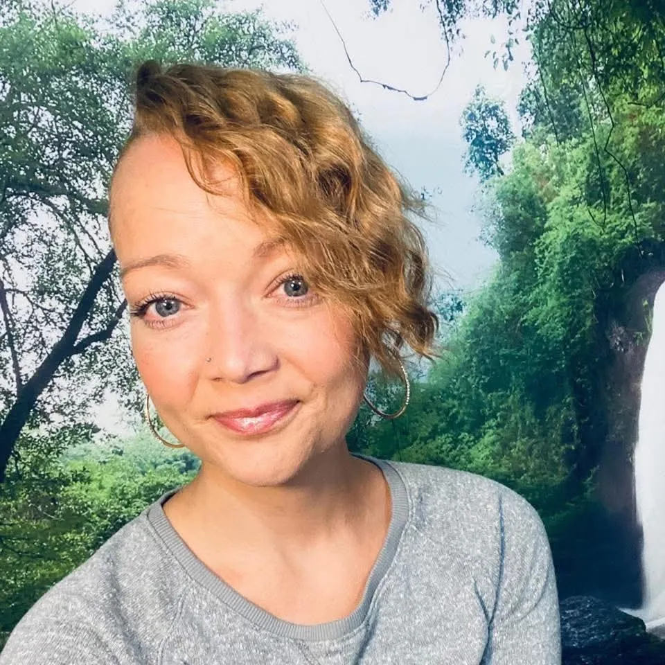 A smiling woman with light brown curly hair and blue eyes, wearing a gray shirt, in front of a backdrop of trees and a waterfall.