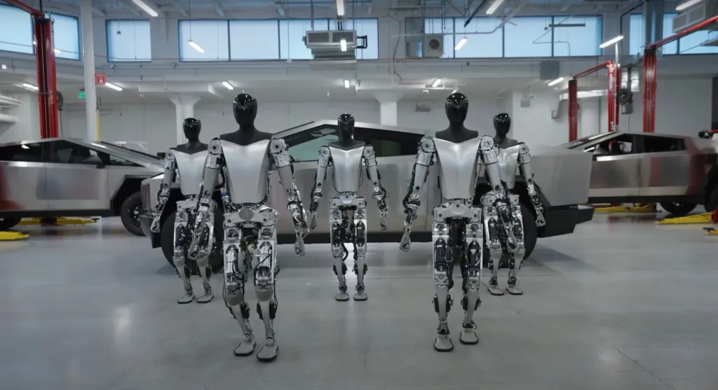 Five Tesla Optimus robots standing in front of two Tesla Cybertrucks in a garage.