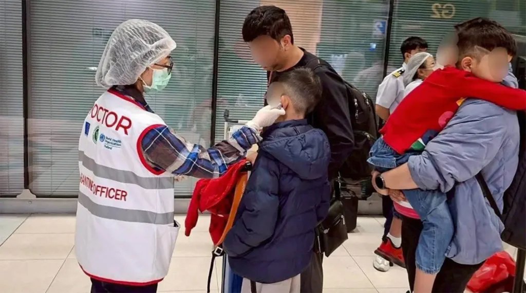 A Thai Department of Disease Control officer in PPE screens a child at an airport.