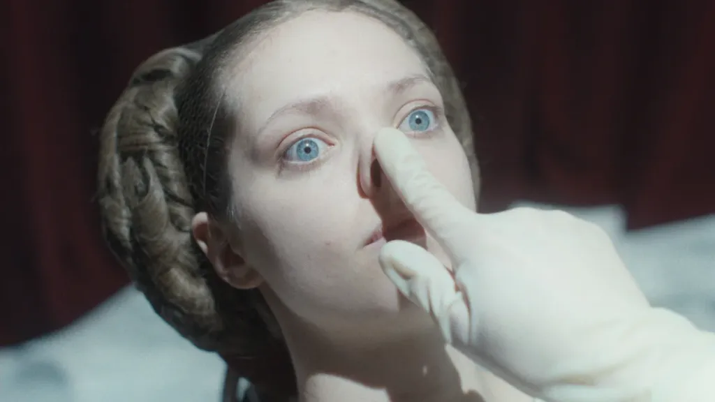 A woman with wide, piercing blue eyes lies back as a gloved hand gently presses on her nose. Her hair is styled in intricate coils on either side of her head, evoking a vintage or theatrical look. The lighting is stark, giving the scene a surreal, clinical tone, while deep red curtains blur in the background.