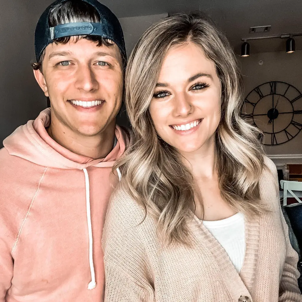 A man wearing a black cap and a pink hoodie standing next to a woman with long blonde hair and a beige cardigan, both smiling.