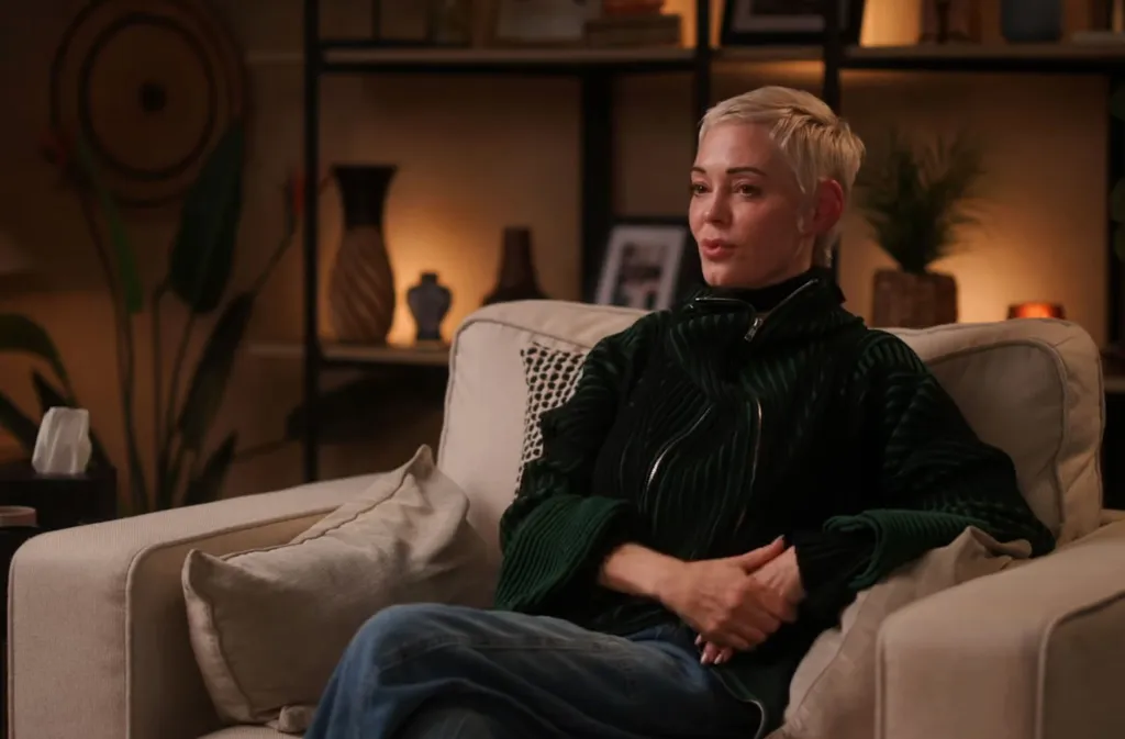 Rose McGowan sits on a beige couch, wearing a green ribbed jacket and blue jeans.
