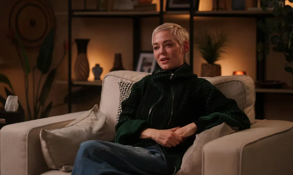 Rose McGowan in a dark green jacket and blue jeans sitting on a light-colored armchair in a cozy room.
