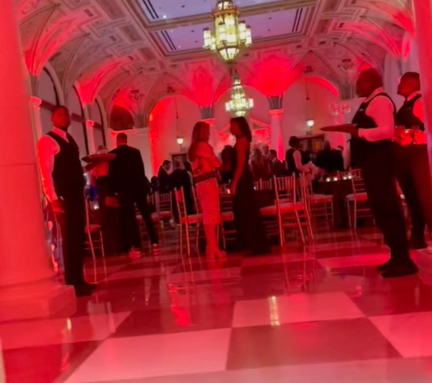 People mingling in a ballroom with red lighting and checkered floors.