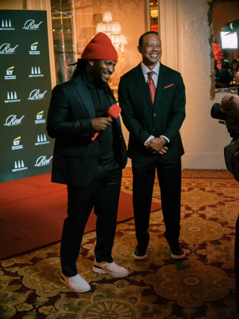 Tiger Woods smiles next to another man in a red knit cap and holding a red microphone, standing in front of a Red and EY branded backdrop.