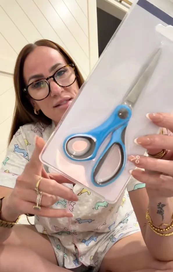 A woman wearing glasses and pajamas patterned with dachshunds holds a pair of blue-handled scissors in a plastic blister pack.