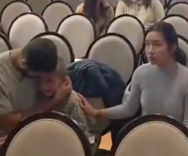 A man comforting a crying child in a room with a woman watching.