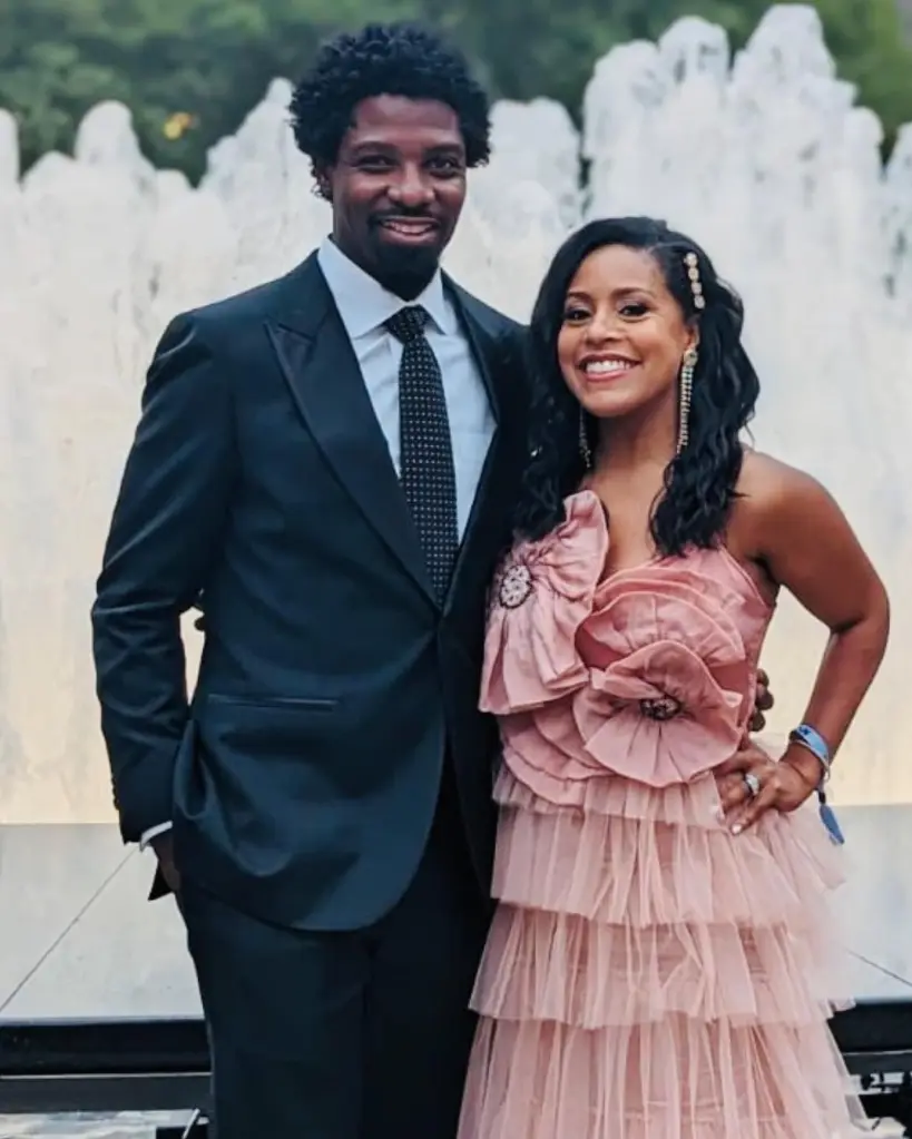 Sheinelle Jones, in a pink strapless dress, and her husband Uche Ojeh, in a blue suit, blue shirt and tie, smiling in front of a fountain.