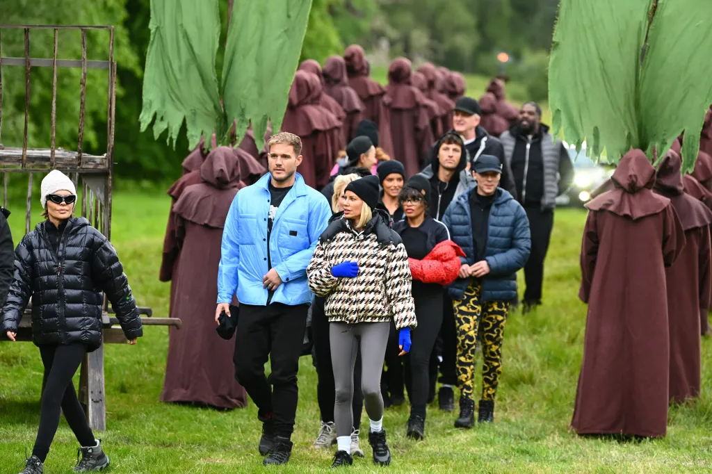 Cast members of The Traitors reality show walking through a field, flanked by people in brown robes.