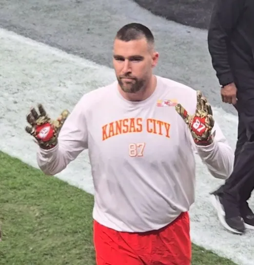 Travis Kelce, wearing a Kansas City shirt and holding up gloved hands, waves to fans.