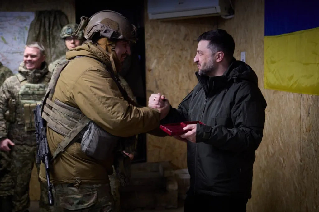 Volodymyr Zelenskyy shaking hands with a soldier while holding an open red box.