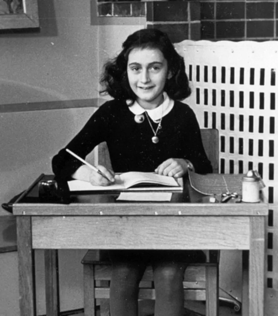 Anne Frank sitting at a desk and writing in a notebook.