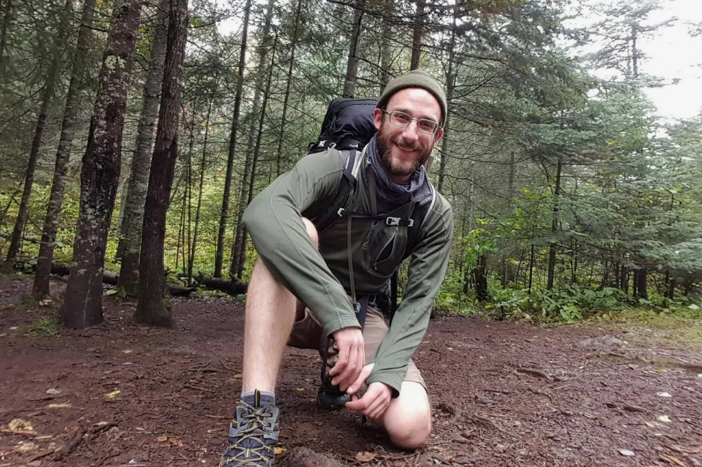 Alex J. Pretti, a man shot by a federal officer, kneels on a dirt path in a forest while wearing hiking gear and smiling at the camera.