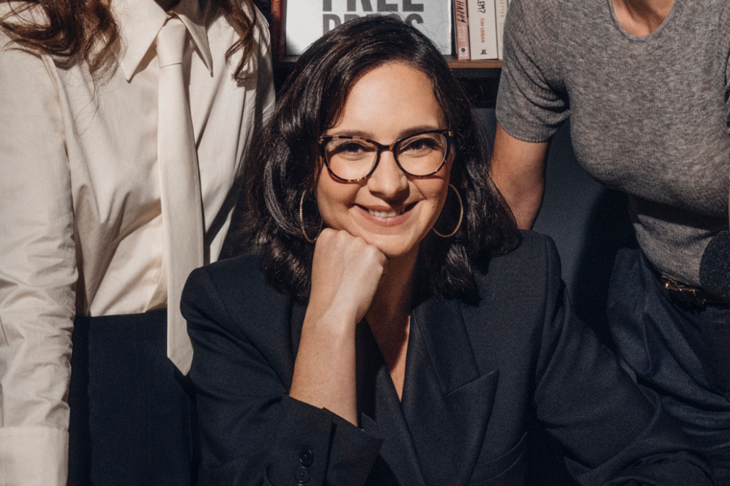 Bari Weiss, co-founder of The Free Press, in a black blazer, miling at the camera with her chin resting on her hand.