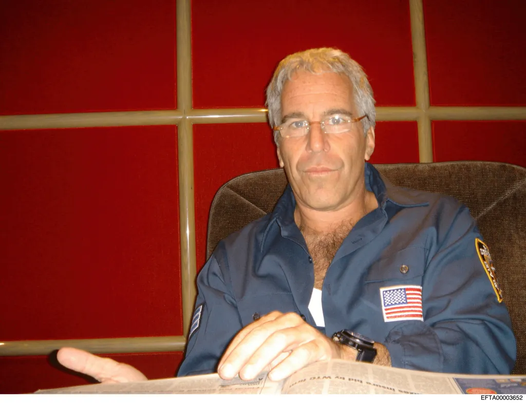Jeffrey Epstein sits in a chair in a dark blue shirt with an American flag patch on the sleeve.