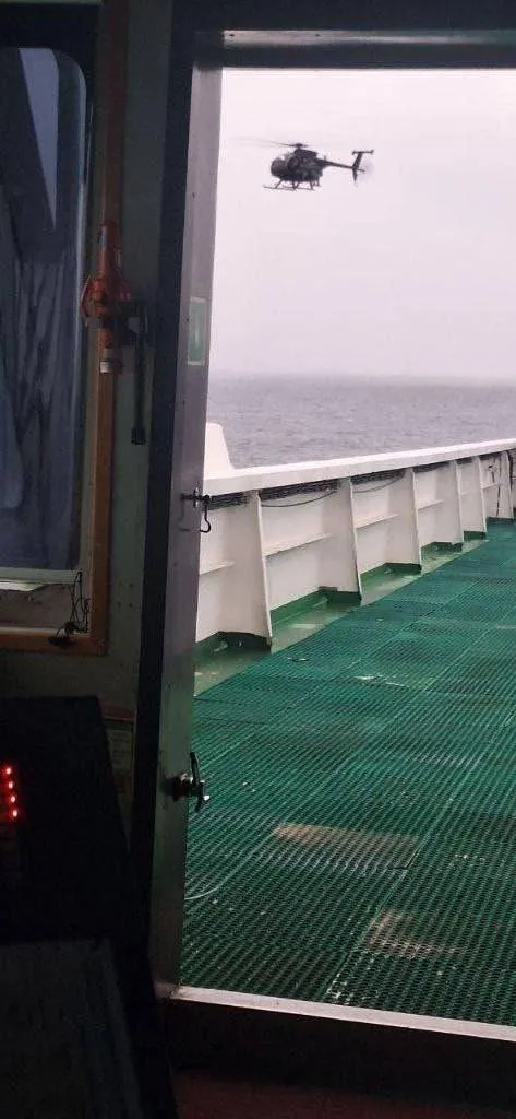A military helicopter hovers over the ocean near the deck of a ship.