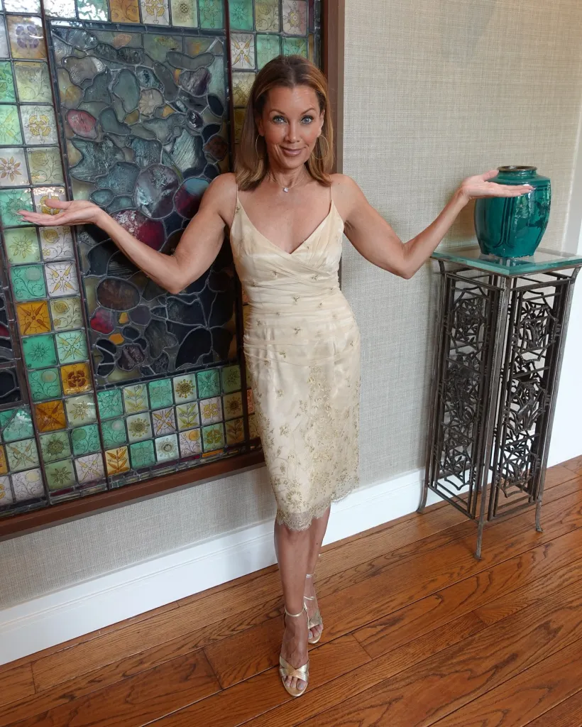 Vanessa Williams in a shimmering champagne dress, with arms outstretched, standing between a stained-glass window and a teal vase on a stand.
