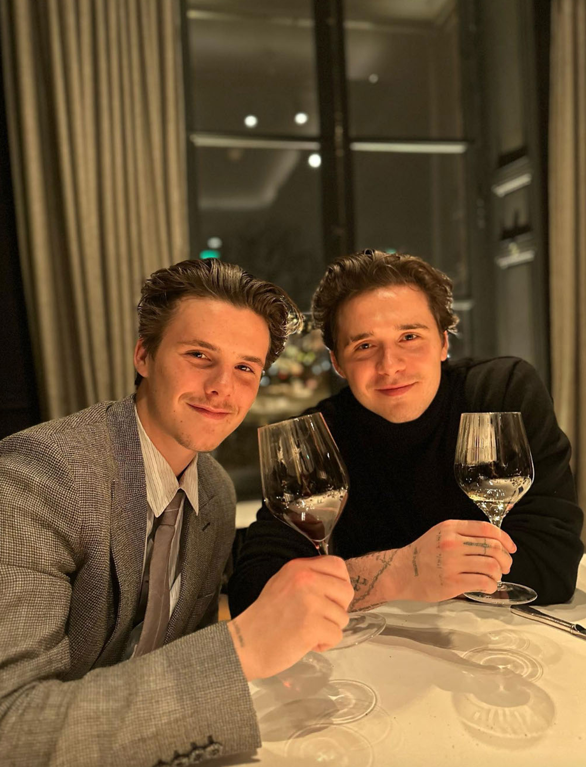 Cruz and Brooklyn Beckham smiling and holding wine glasses at a dinner table.