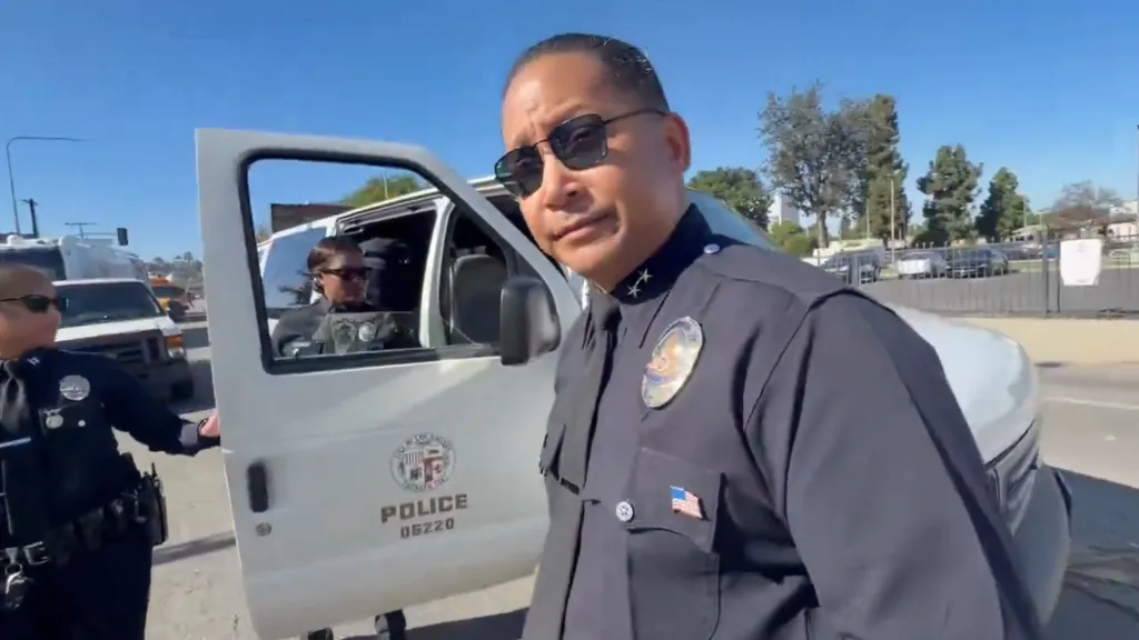 LAPD Deputy Chief Alan Hamilton in uniform and sunglasses looking at the camera, standing next to a police van with an open door.