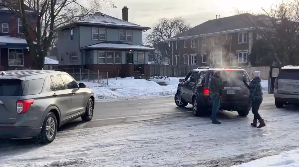 Two people in winter clothing stand between two dark SUVs on a snowy residential street in Minneapolis.