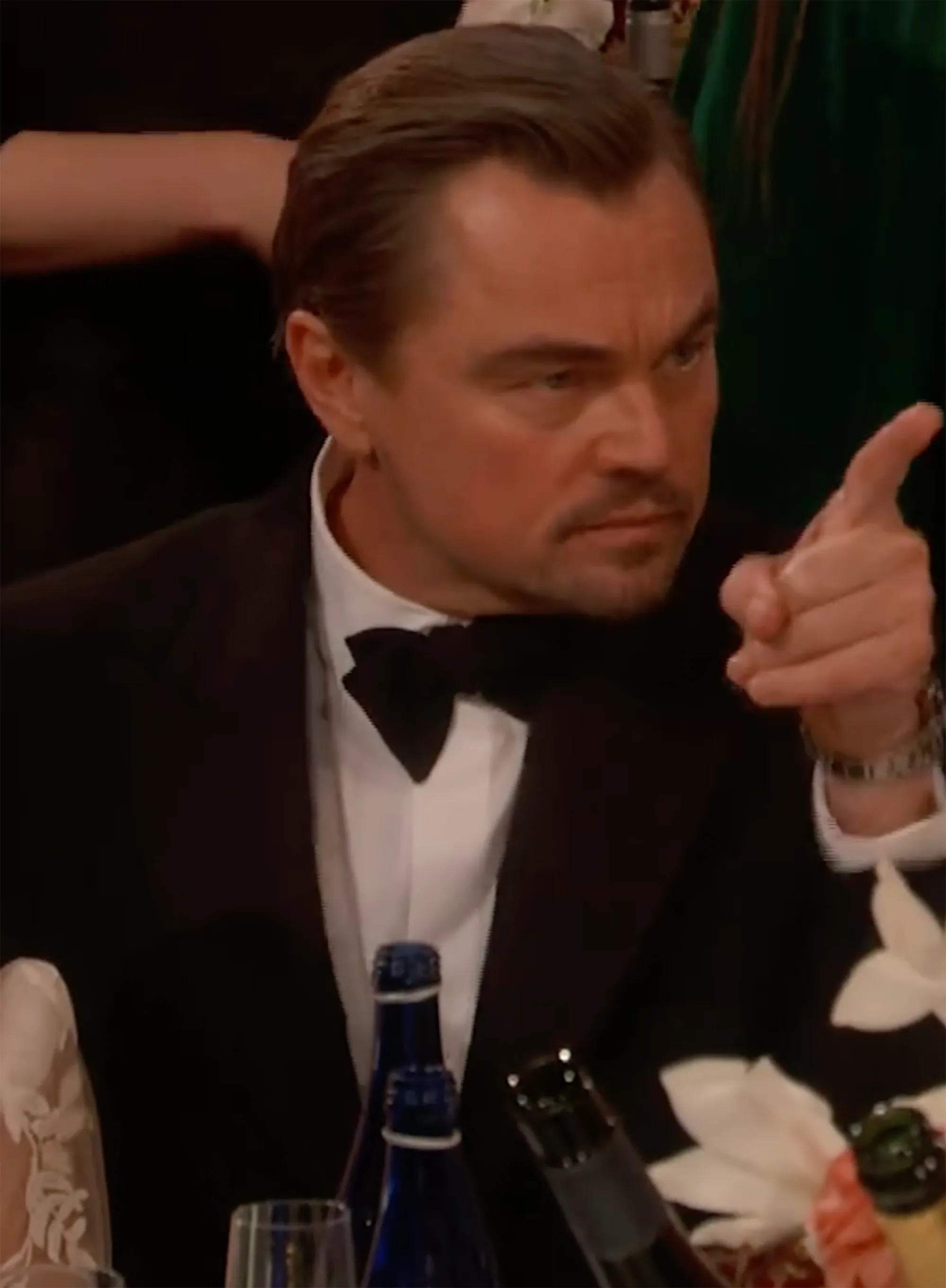 Leonardo DiCaprio, wearing a black suit and bow tie, points his finger while looking forward.