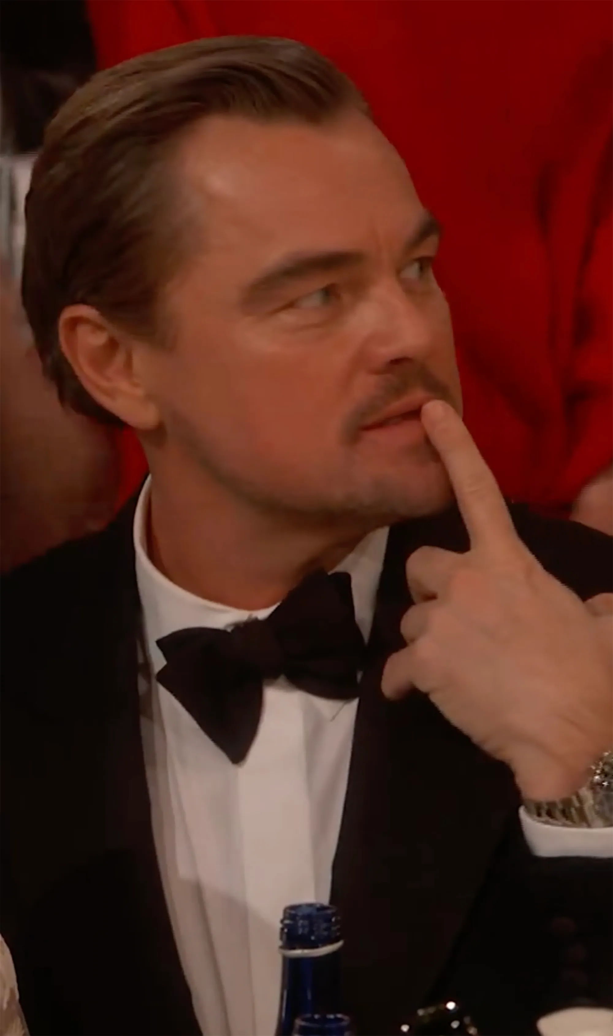 Leonardo DiCaprio in a tuxedo, with a finger touching his upper lip and a bottle on the table in front of him.