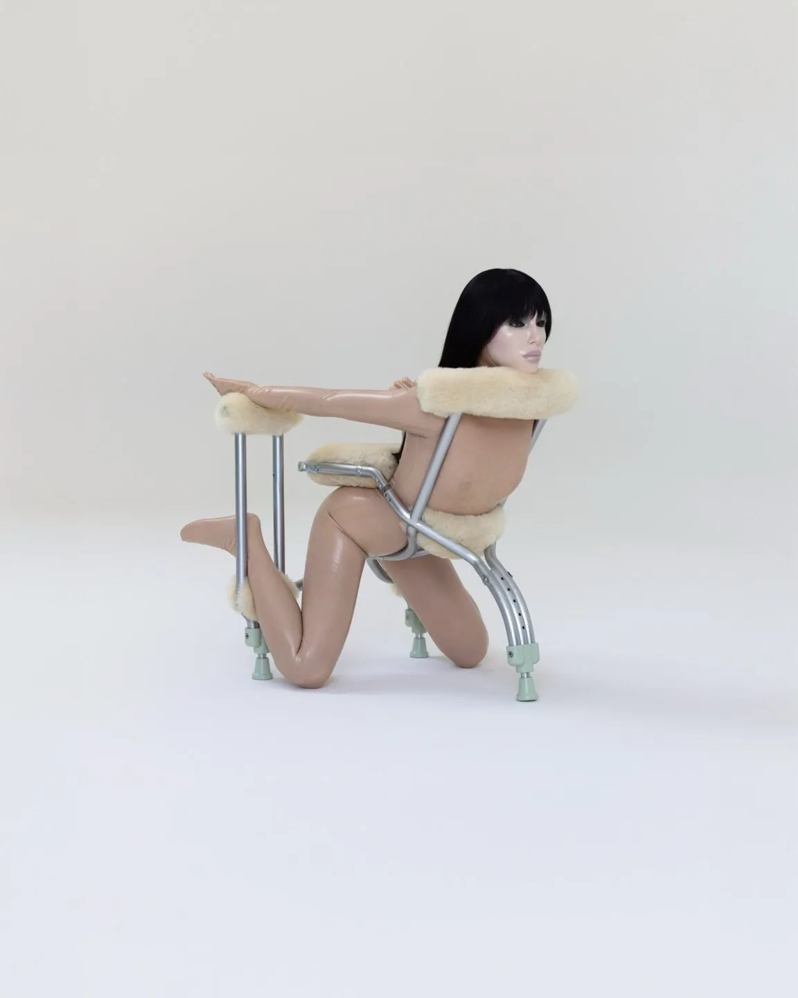 A nude mannequin, with long black hair, kneeling and propping itself up on metal crutches, with cream-colored furry padding.