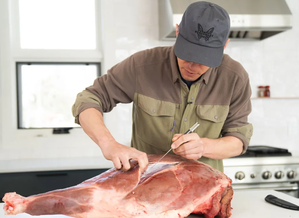 Man in a cap using a knife to butcher a large piece of meat in a kitchen.