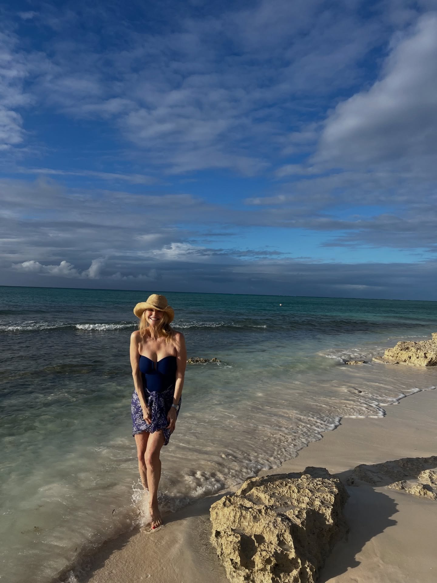 Christie Brinkley in a navy swimsuit and straw hat, wading in the ocean at a beach.