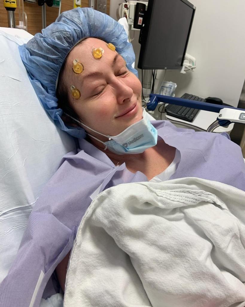 Shannen Doherty in a hospital bed with electrodes on her forehead, wearing a surgical cap and a face mask under her chin.