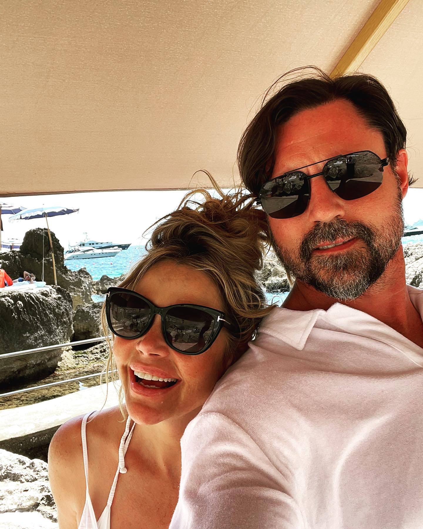 Denise Richards and Aaron Phypers smiling under an awning by the sea.