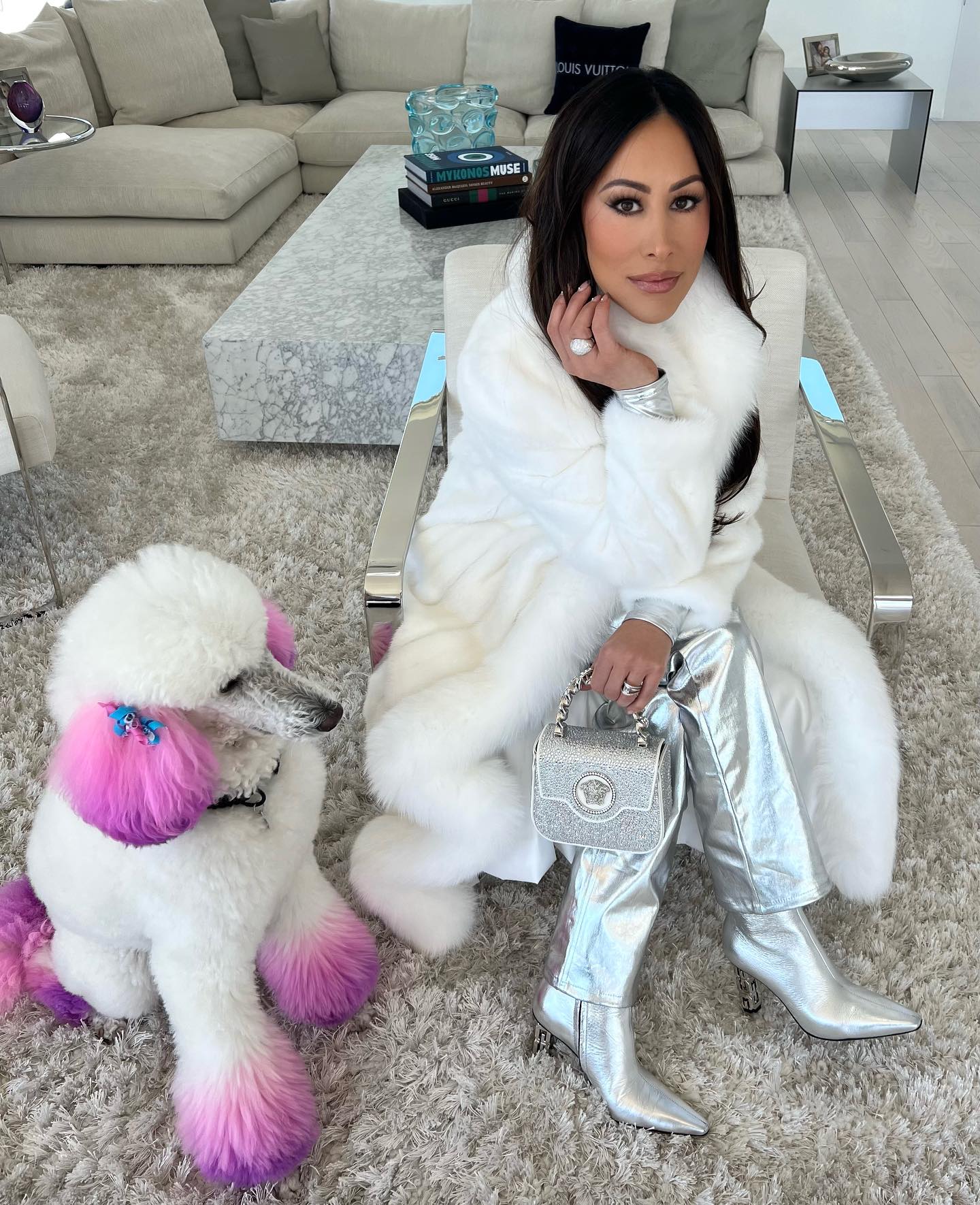 Angie Katsanevas with her poodle Celia, wearing a white fur coat, silver pants, and silver boots, holding a sparkly silver handbag.
