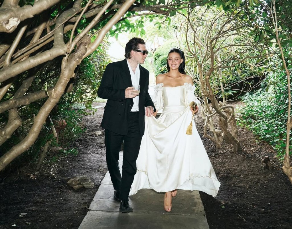 Brooklyn Beckham, in a black suit and white shirt, and Nicola Peltz, in a cream wedding gown, walking on a path surrounded by trees and bushes.