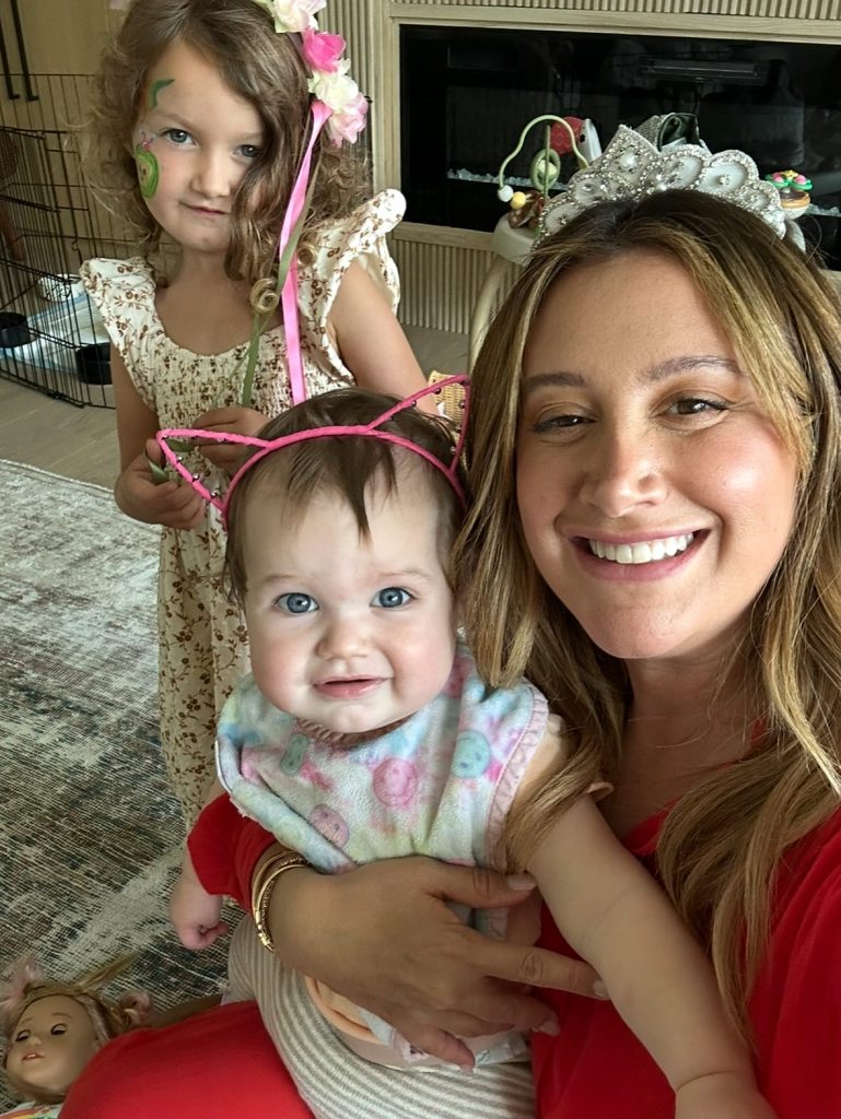 Ashley Tisdale holding a baby with a cat ear headband, and an older child wearing a floral dress and flower headband.