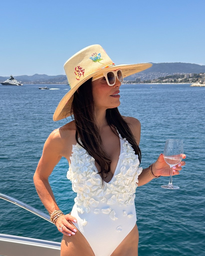 Bethenny Frankel in a white swimsuit on a yacht with a drink in her hand.