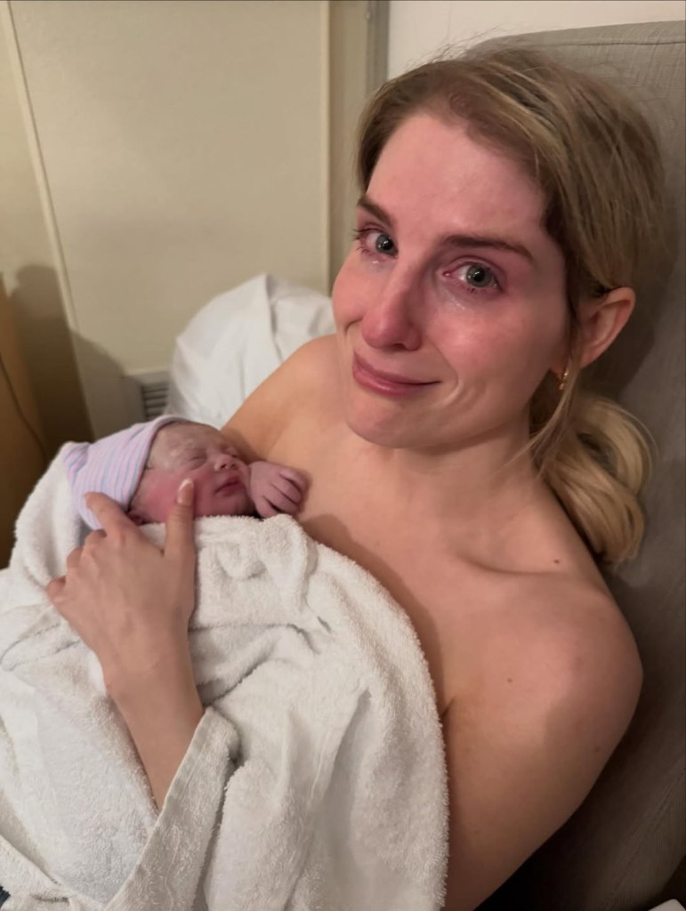 Meghan Trainor with teary eyes holding a newborn baby wrapped in a white towel.