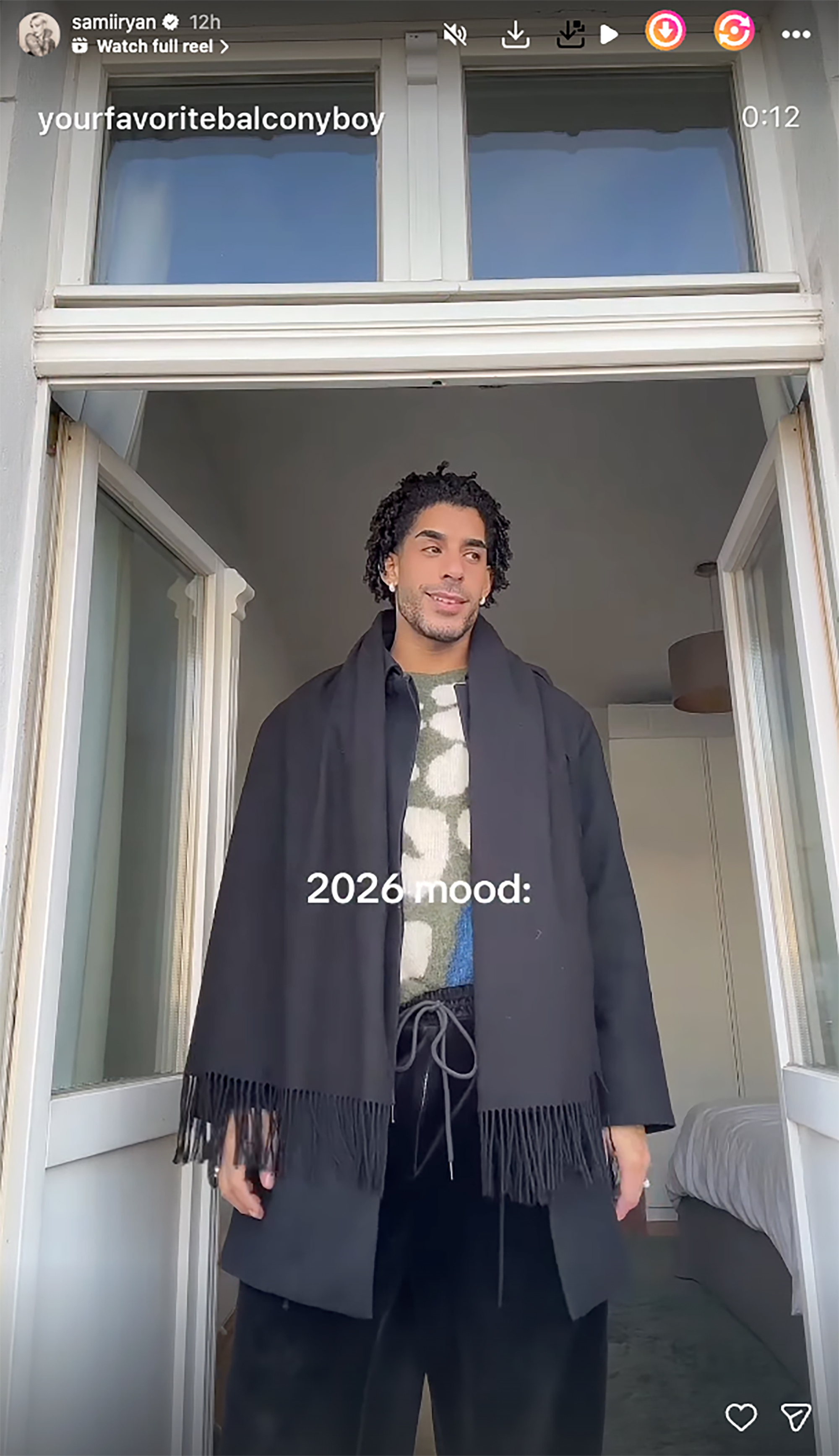 A person wearing a black jacket and scarf with a patterned sweater and black pants stands in a doorway, smiling. Text says 