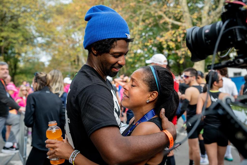Sheinelle Jones, wearing a 2023 finisher's medal, in a black tank top, embraces her husband Uche Ojeh, in a black t-shirt and blue hat, holding a bottle of orange sports drink.