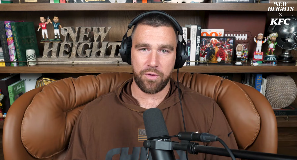 Travis Kelce in headphones and a brown hoodie, sitting in a leather chair in front of a bookshelf with 