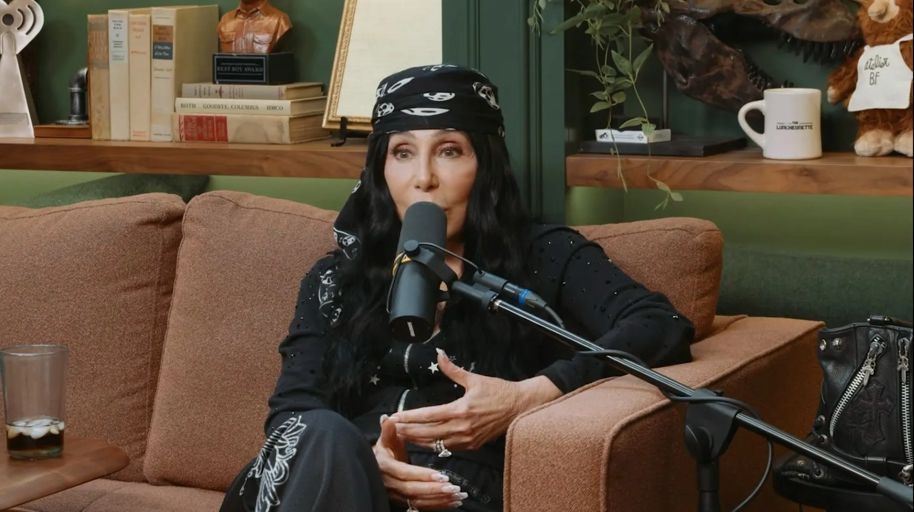 Cher talking into a microphone while sitting on a brown couch.