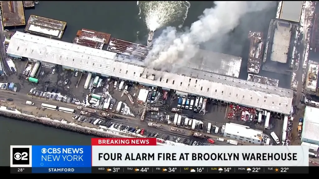 Aerial view of a four-alarm warehouse fire in Brooklyn, with a fire boat spraying water on the burning structure.