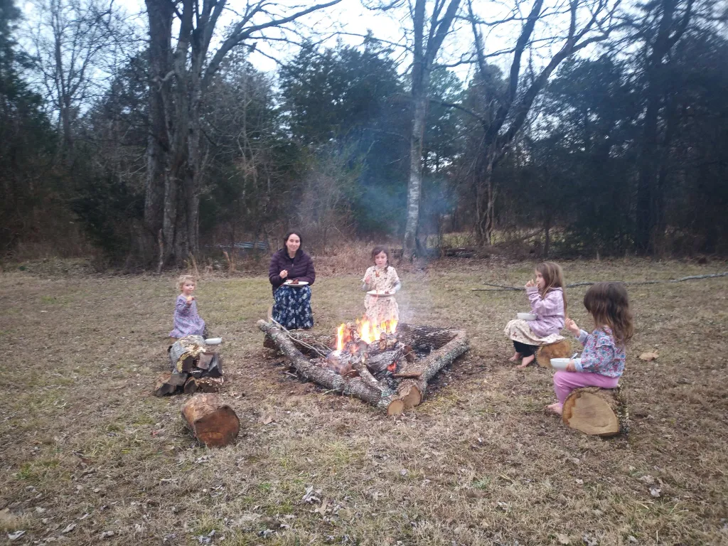 A woman and four young children sit around a campfire in a grassy clearing, holding plates and bowls.