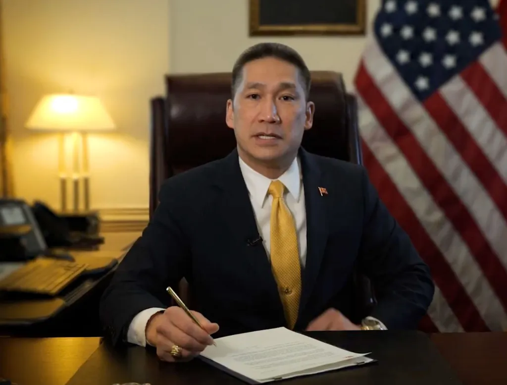 Hung Cao, a man in a suit and tie, signing documents at a desk with an American flag in the background.