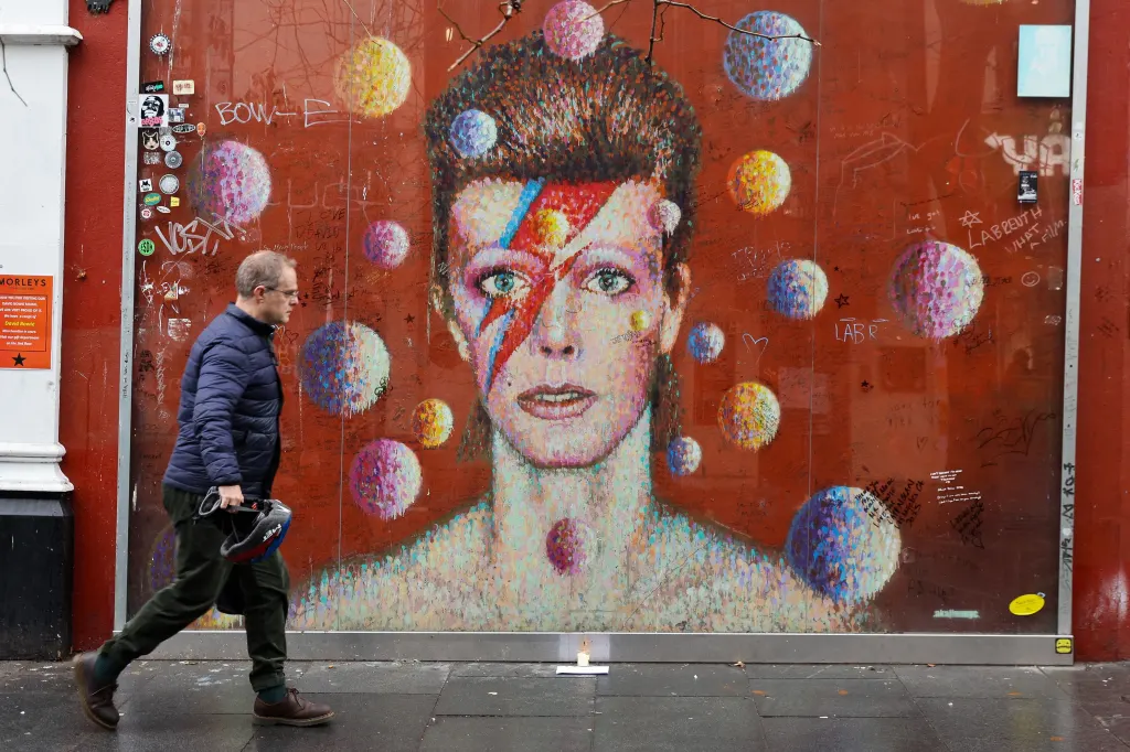 A man walks past a mural of David Bowie with a lightning bolt painted across his face and colorful planets floating around him.