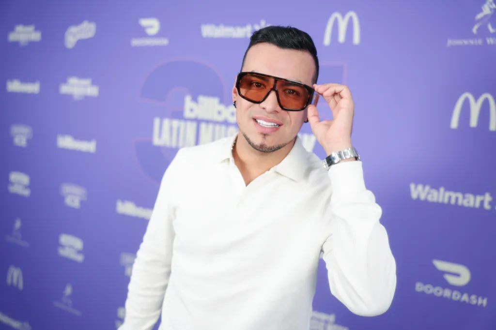 Yeison Jimenez adjusting his sunglasses, posing in front of a purple backdrop with