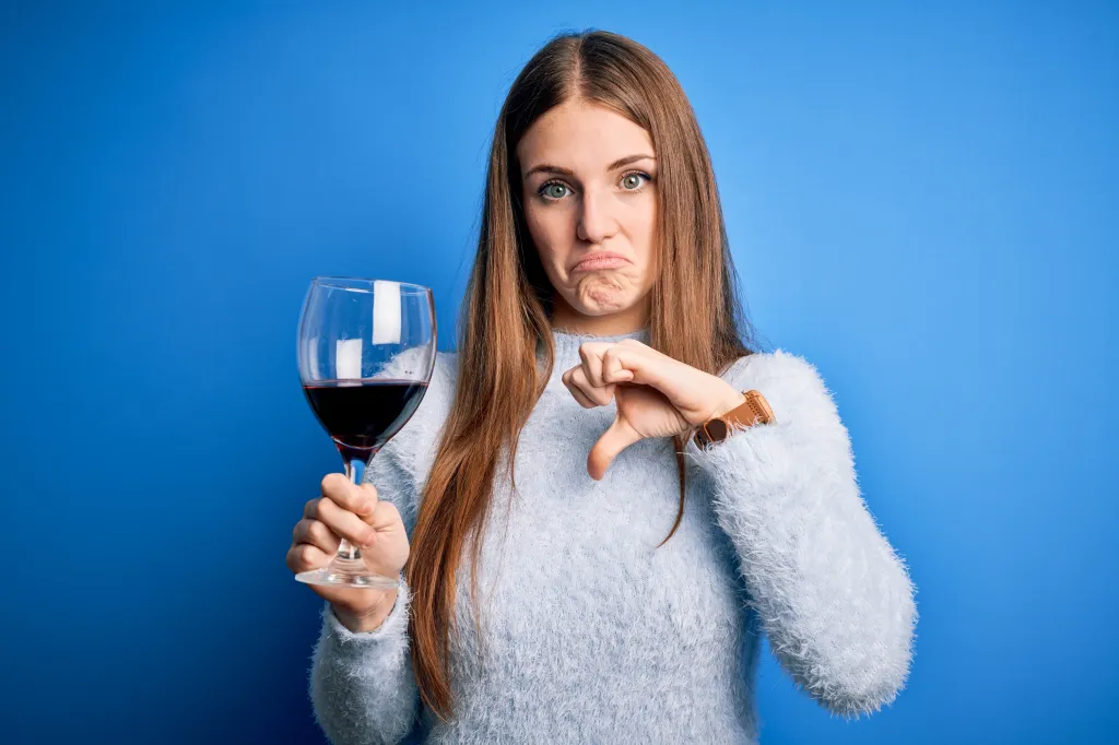 A displeased woman holding a glass of red wine gives a thumbs down.