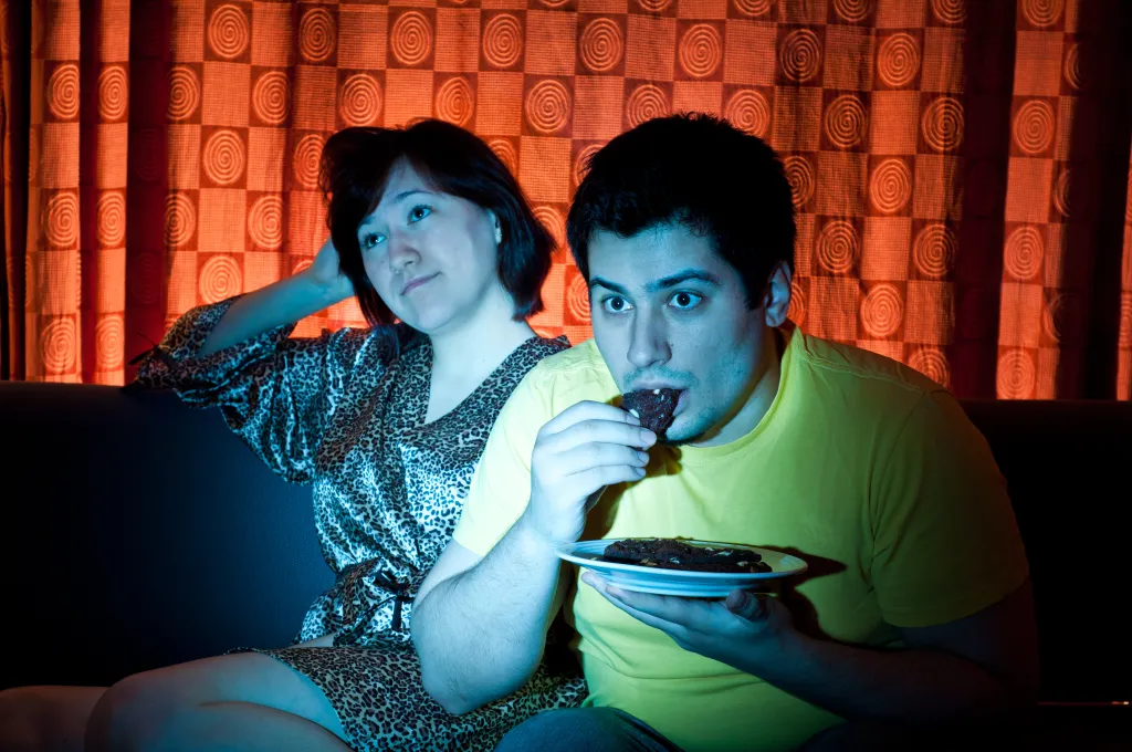 A man eating a cookie and looking intently at a TV screen, while a woman beside him looks bored.