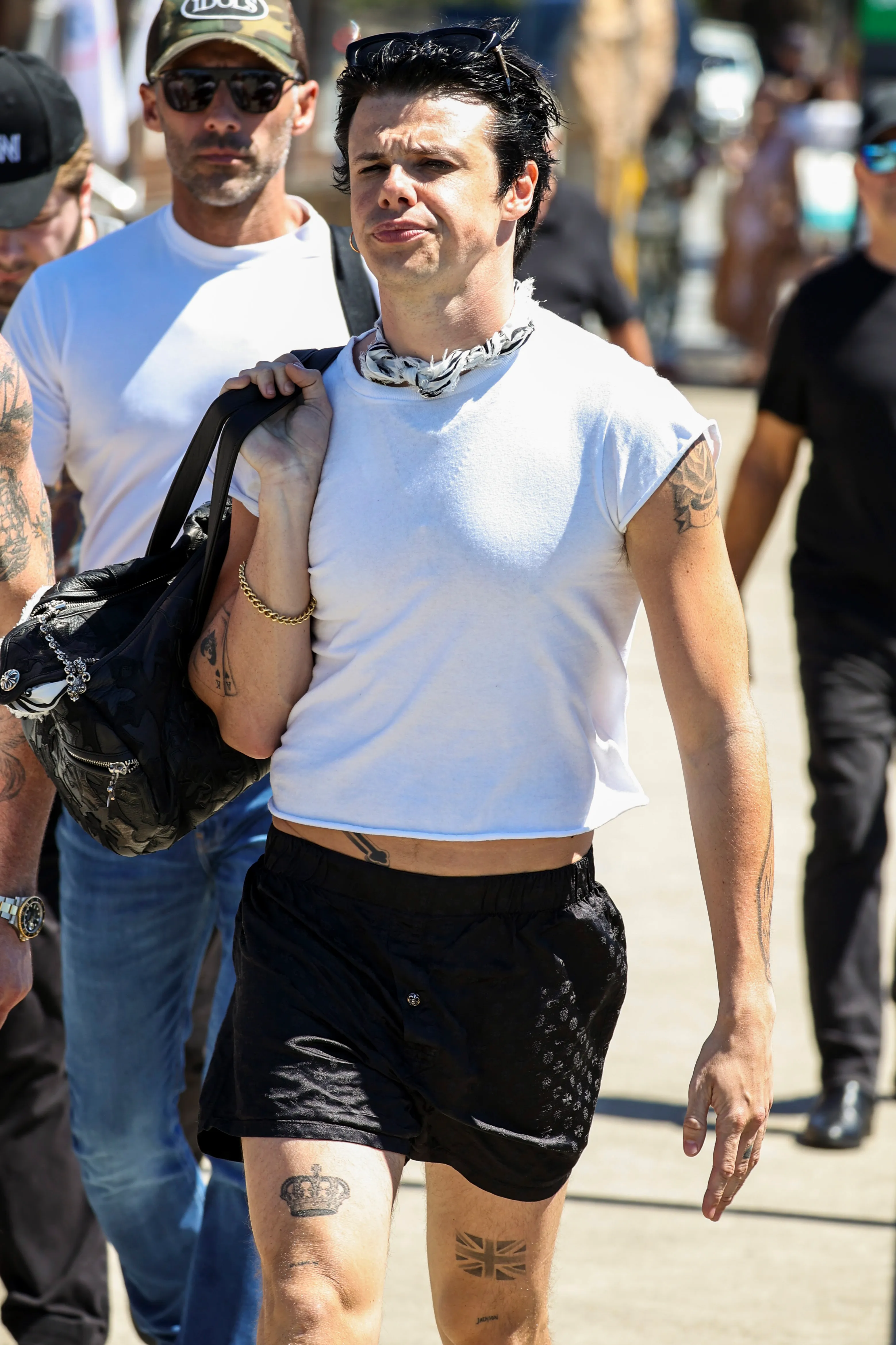 Yungblud, wearing a white sleeveless top and black shorts, carries a duffel bag while walking.