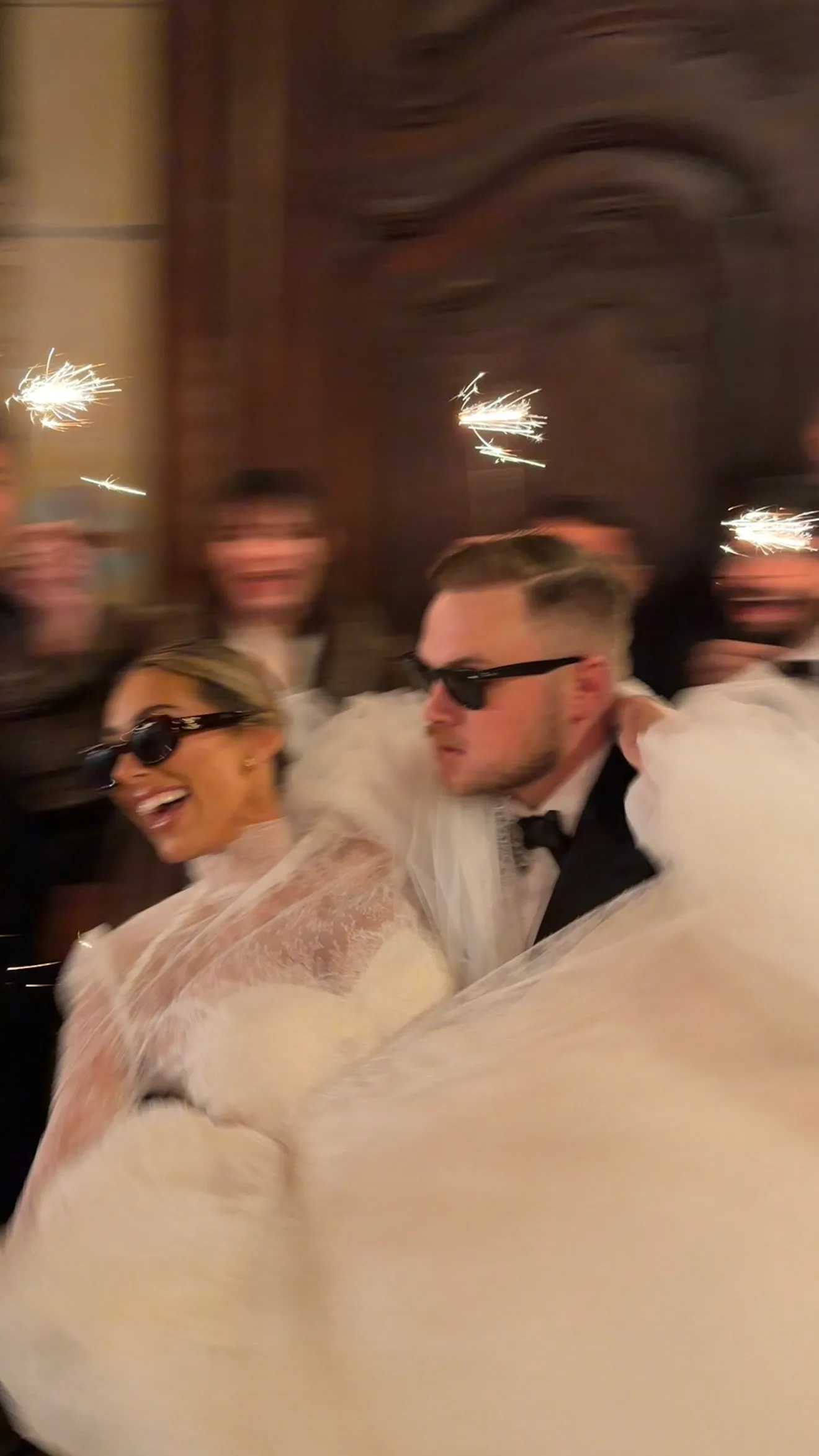 Zach Bryan carrying Samantha Leonard, both in sunglasses, with sparklers in the background.