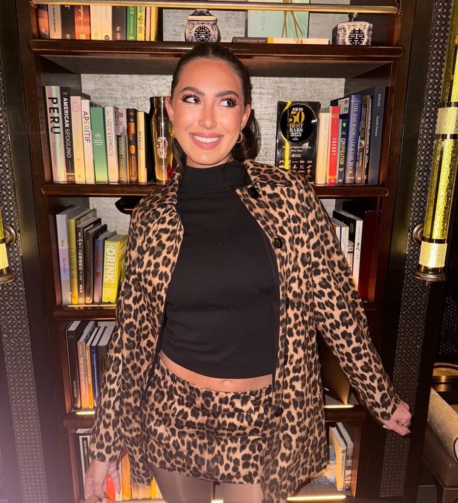 Samantha Leonard, wearing a leopard print jacket and skirt, smiles and looks off to the side, standing in front of a bookshelf.